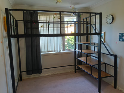 A black metal loft bed frame with built-in wooden steps and shelves, set up in a room with beige walls and a window.